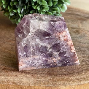 May include: A faceted amethyst crystal with shades of purple, white, and reddish-brown. The crystal is a rectangular shape with a polished surface, showcasing its natural crystalline structure. The background includes a green plant and a wooden surface.