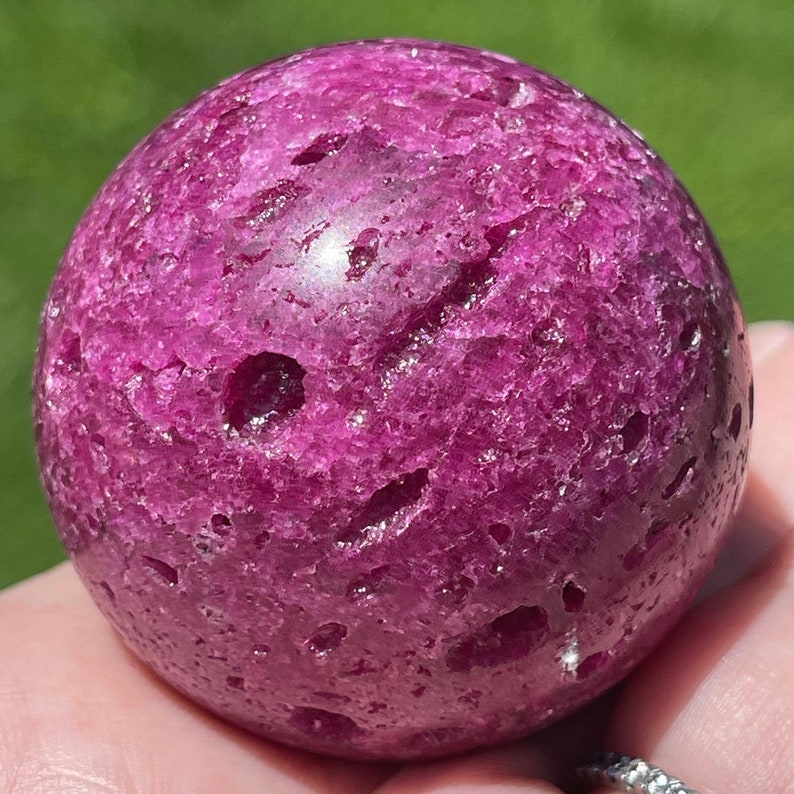 May include: A close-up of a round, deep magenta-colored sphere with a textured surface. The sphere has several small, irregular holes and a slightly uneven shape. The background is blurred green.