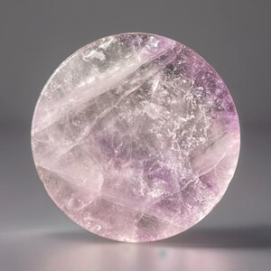 May include: A round, polished amethyst stone with a mix of light purple and white hues. The stone has a translucent quality, with visible internal patterns and variations in color. The overall shape is a smooth, circular disc.
