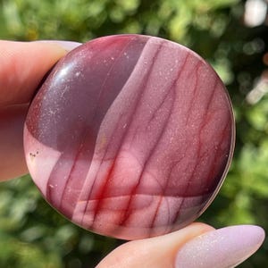 May include: A polished, round stone with a mix of burgundy, red, and pink hues. The stone has a smooth, glossy surface with natural veining patterns. The colors blend together, creating a unique and organic appearance. The stone is held in a hand.
