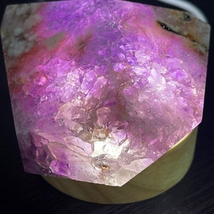 May include: A large, faceted amethyst crystal illuminated from within, showcasing shades of purple and pink. The crystal sits on a wooden base, creating a glowing effect. The rough texture of the crystal is visible.
