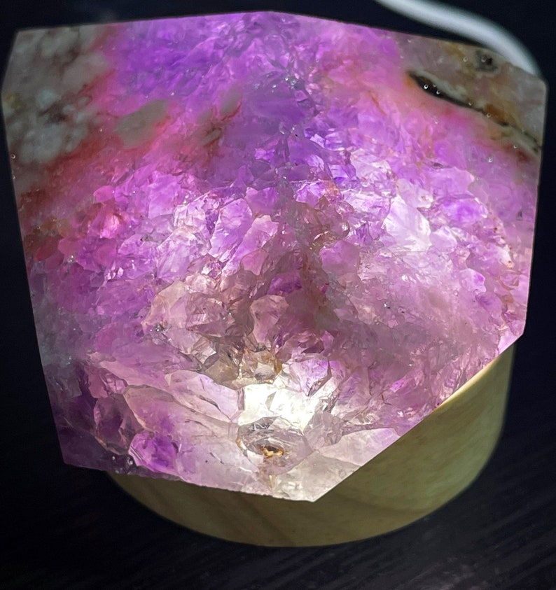 May include: A large, faceted amethyst crystal illuminated from within, showcasing shades of purple and pink. The crystal sits on a wooden base, creating a glowing effect. The rough texture of the crystal is visible.