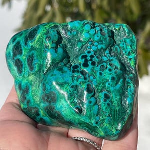 May include: A vibrant, natural malachite stone with a rich blend of green and teal hues. The stone's surface features a textured pattern with dark green and black spots, creating a unique and eye-catching appearance. The stone is a natural mineral specimen.