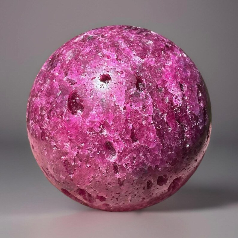 May include: A spherical, textured gemstone in shades of pink and magenta. The surface has a rough, pitted appearance, with variations in color intensity. The gemstone is a solid, opaque sphere, likely a decorative item or a mineral specimen.