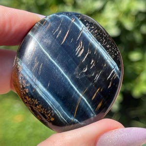 May include: A polished, oval-shaped blue tiger's eye gemstone. The stone features bands of blue, gold, and brown, with a glossy finish. The gemstone is held in a hand against a blurred green background.