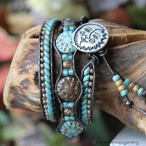 May include: A multi-strand leather wrap bracelet with turquoise and brown beads, featuring a silver charm with a face on it. The bracelet is wrapped around a piece of driftwood.