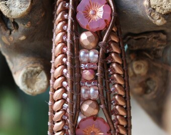 May include: A multi-strand leather bracelet with copper beads, pink glass beads, and pink flower-shaped charms. The bracelet is wrapped around a piece of driftwood.