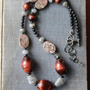 May include: A handcrafted necklace featuring a mix of natural stone beads. The necklace includes oval and round beads in shades of brown, gray, and black. The clasp is silver-toned. The necklace is displayed on a blue-gray surface.