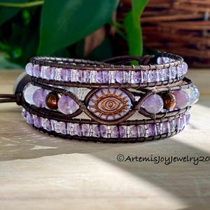 May include: Wrap bracelet with purple and clear beads, brown leather, and a central eye design. The bracelet is multi-layered and features a mix of bead sizes and textures. The design includes the text "Artemisjoyjewelry2025".