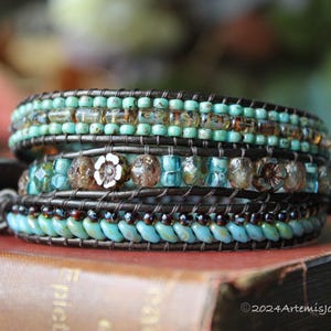 May include: A multi-strand wrap bracelet with turquoise, brown, and clear beads. The bracelet features a leather band and a silver-toned flower charm. The beads are arranged in rows, creating a textured and layered effect. The bracelet is displayed on a book.