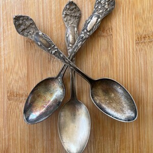 May include: Three silver spoons with ornate floral designs on the handles. The spoons are arranged in a star shape on a wooden surface.