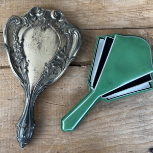 May include: Two vintage hand mirrors. One is silver with an ornate design and the other is green with black and white accents.