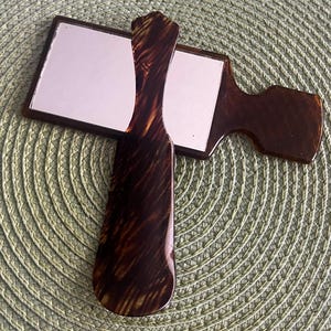 May include: A handheld mirror and a matching hairbrush with a dark brown faux tortoiseshell pattern. The mirror has a rectangular reflective surface with rounded corners, and the hairbrush features a long, slender handle and a wide head.