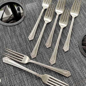 May include: A collection of silver forks with ornate floral detailing on the handles. The forks are arranged on a textured gray surface, showcasing their elegant design. The tines are clearly visible, highlighting the silverware's functionality.
