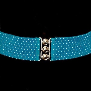 May include: A teal polka dot belt with a silver buckle. The belt is wide and elastic, with small white polka dots. The buckle has three silver buttons. The belt is shown on a black background.