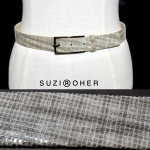 May include: A neutral-toned belt with a textured pattern and a rectangular buckle. The belt is displayed on a mannequin and features the brand name "SUZI® OHER". The belt's design is visible in a close-up shot.