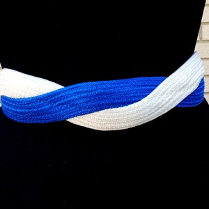 May include: A braided belt in a striking combination of white and royal blue. The belt is wrapped around a black form, showcasing its woven texture and contrasting colors. This accessory adds a pop of color and style.