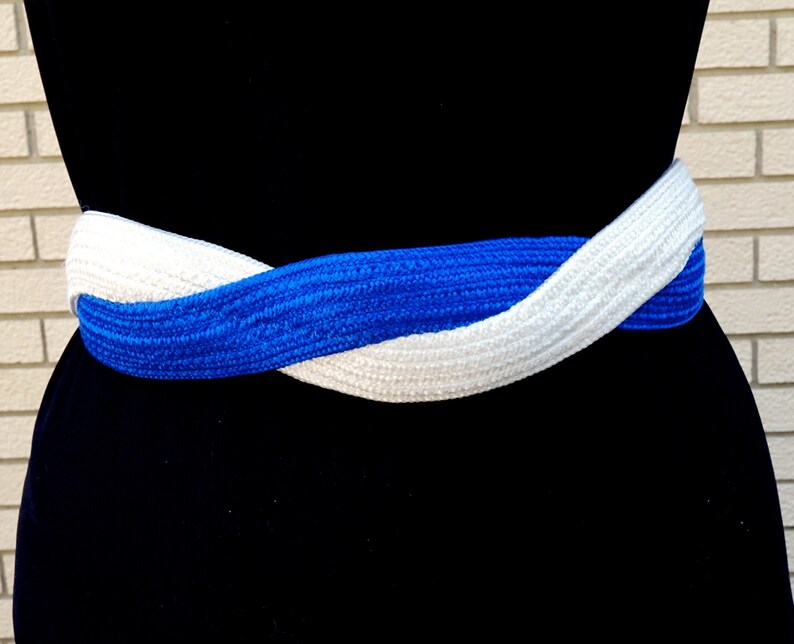 May include: A braided belt in a striking combination of white and royal blue. The belt is wrapped around a black form, showcasing its woven texture and contrasting colors. This accessory adds a pop of color and style.