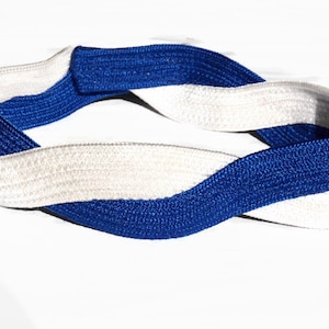 May include: A braided headband in a twisted design, featuring alternating bands of white and royal blue. The headband is made of a woven material and is shown against a white background.