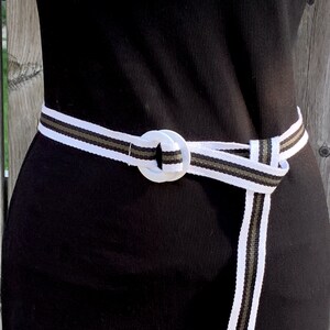 May include: A white, olive green, and black striped belt with a white circular buckle is wrapped around a black dress. The belt is knotted at the waist. The background is a wooden fence.