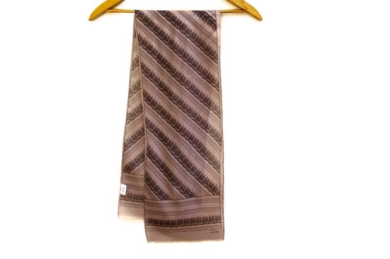 Puede incluir: Bufanda color topo con un estampado de rayas diagonales de dise&ntilde;os florales marr&oacute;n oscuro. La bufanda est&aacute; colgada de una percha de madera sobre un fondo blanco. La bufanda tiene forma rectangular.