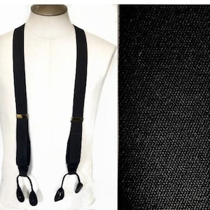 May include: Black suspenders with brass-coloured hardware and button attachments. The suspenders are displayed on a white mannequin. A close-up of the black fabric shows a textured weave.
