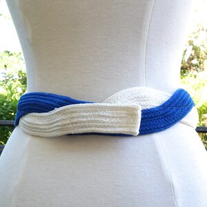 May include: A woven belt in a twisted design, featuring alternating sections of bright blue and off-white. The belt is displayed on a white mannequin, showcasing its texture and style. The belt is a fashion accessory.
