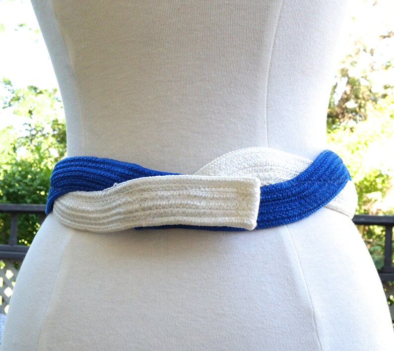 May include: A woven belt in a twisted design, featuring alternating sections of bright blue and off-white. The belt is displayed on a white mannequin, showcasing its texture and style. The belt is a fashion accessory.