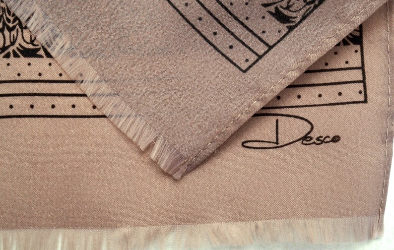 Puede incluir: Primer plano de una bufanda color malva con borde deshilachado. La bufanda presenta un dise&ntilde;o geom&eacute;trico negro y la palabra "Desco" en cursiva. La tela parece suave y tiene un brillo sutil.