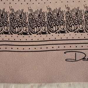 Puede incluir: Bufanda beige con un dise&ntilde;o de espigas de trigo negras en el borde superior. El dise&ntilde;o incluye un borde punteado y la firma "Desco". La bufanda tiene un borde con flecos. Aproximadamente 100 cm de largo.