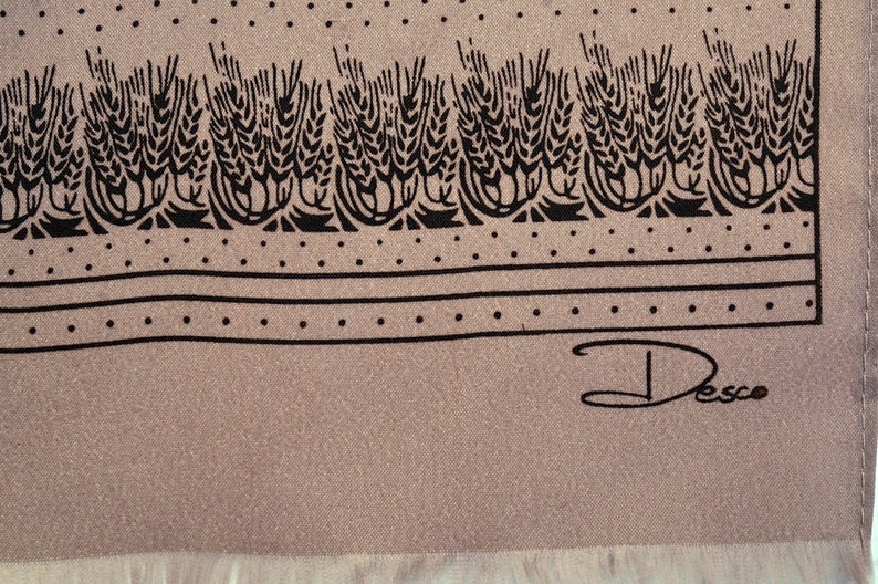 Puede incluir: Bufanda beige con un dise&ntilde;o de espigas de trigo negras en el borde superior. El dise&ntilde;o incluye un borde punteado y la firma "Desco". La bufanda tiene un borde con flecos. Aproximadamente 100 cm de largo.