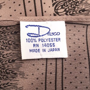Puede incluir: Primer plano de una tela con un patr&oacute;n de trigo negro sobre un fondo color topo. Una etiqueta blanca con texto azul dice "Desco 100% Polyester RN 14055 Made in Japan."