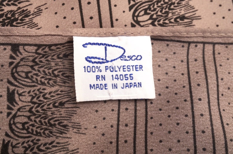 Puede incluir: Primer plano de una tela con un patr&oacute;n de trigo negro sobre un fondo color topo. Una etiqueta blanca con texto azul dice "Desco 100% Polyester RN 14055 Made in Japan."