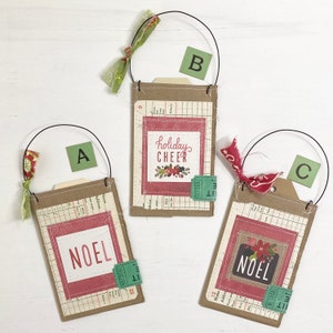 May include: Three rectangular brown cardstock tags with wire hangers and fabric ties. Each tag features a vintage ledger paper background, a red square frame, and a green ticket stub. Tag A says NOEL, B says holiday CHEER, and C says NOEL.
