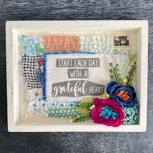 May include: A white rectangular frame containing a fabric collage of various patterned patches. A central cream-colored fabric piece features the text "START EACH DAY WITH A GRATEFUL HEART." Felt flowers in blue, magenta, and green accent the corner.