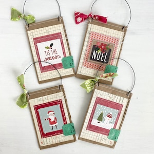 May include: Four rectangular hanging ornaments made of brown cardstock with grid paper backgrounds. Each features a central holiday-themed graphic, a green raffle ticket accent, and a wire hanger with a fabric bow. Designs include text reading TIS THE SEASON and NOEL, a Santa Claus illustration, and a snowman.