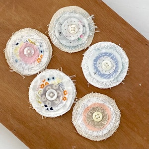 May include: Five handmade fabric flower embellishments. Each circular piece features layered fabric in various patterns and colors, with a button center. The edges are frayed, giving a rustic, vintage aesthetic. The color palette includes white, pink, blue, and neutral tones.