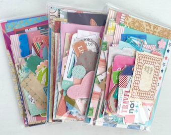 May include: Three packs of colourful paper scraps for scrapbooking and crafting. The packs are filled with patterned paper, stickers, and embellishments. The paper scraps are in various colours, including pink, blue, green, and yellow. The packs are labelled with the words "Play, smile, laugh" and "Travel, not worry".
