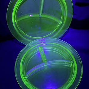Op de afbeelding: Twee ronde, verdeelde borden in een levendige neon groene tint. Elk bord is verdeeld in drie compartimenten, met een opstaande rand en een centrale verdeler. De borden zijn gemaakt van glas en worden verlicht met zwart licht, waardoor een gloeiend effect ontstaat.