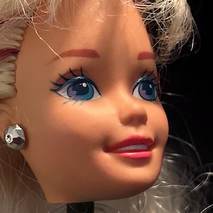 Puede incluir: Primer plano de la cabeza de una muñeca con cabello rubio y ojos azules. La muñeca tiene labios rosados y un pendiente plateado facetado. La cara de la muñeca está ligeramente girada hacia un lado, con una expresión neutra.