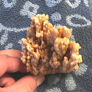 May include: A light brown, textured mineral specimen with a crystalline structure. The mineral is held by a hand, showcasing its intricate, layered formations. The color is a warm, earthy tone, with a rough, uneven surface.