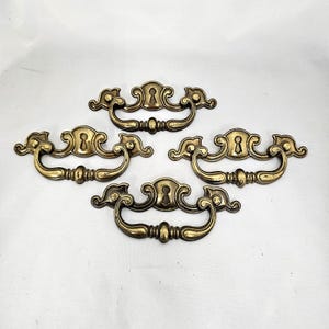 May include: Set of four antique brass drawer pulls. Each pull features a keyhole and a curved handle with decorative detailing. The pulls are arranged on a white background, showcasing their aged, golden finish. Ideal for vintage furniture restoration.