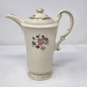 May include: A cream-colored porcelain coffee pot featuring a floral bouquet in shades of pink, blue, yellow, and green. The pot includes a lid with a finial, a curved handle, and a spout, all accented with gold trim and a geometric pattern.