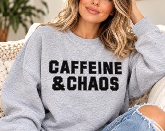 May include: A light gray crewneck sweatshirt featuring the text CAFFEINE & CHAOS printed in bold black block letters across the chest. The garment has long sleeves and a ribbed neckline, cuffs, and hem, providing a classic and comfortable fit.