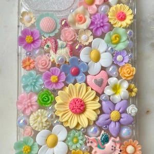 May include: A clear phone case decorated with colorful flowers, glitter, and unicorn charms. The case is filled with a variety of pastel-colored flowers, including daisies, roses, and other floral designs. The case also features small, sparkly unicorn charms.