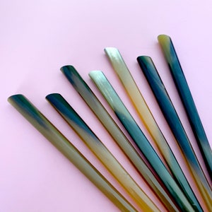 Puede incluir: Conjunto de seis pajitas para beber de cuerno en varios tonos de marrón, verde y azul. Las pajitas están dispuestas en forma de abanico sobre un fondo rosa.