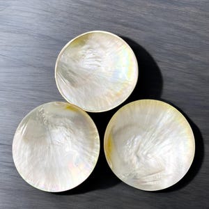 Puede incluir: Tres cuencos blancos de madreperla con brillo iridiscente. Los cuencos son redondos y poco profundos, y están dispuestos en un patrón triangular.