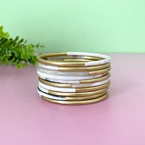 Puede incluir: Una pila de brazaletes blancos y dorados. Los brazaletes están hechos de un material duro y liso y tienen un acabado brillante. La pintura dorada se aplica a la mitad superior de cada brazalete, creando un efecto rayado.