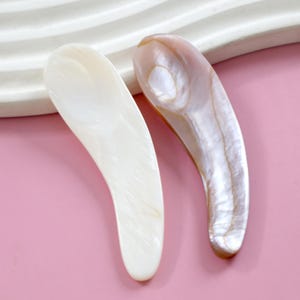 Puede incluir: Dos herramientas de masaje facial de nácar blanco y rosa. Las herramientas tienen forma de cuchara con un mango curvo.