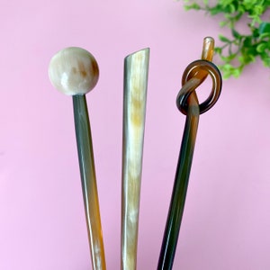 Può includere: Tre bastoncini per capelli marroni e beige. Un bastoncino ha una parte superiore rotonda, liscia e beige. Un bastoncino ha un nodo al centro. Un bastoncino è dritto e ha una punta affilata.